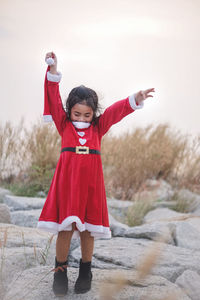 Girl wearing red dress with arms raised standing on rocks against sky