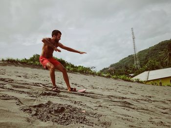 Full length of shirtless man standing on surfboard at beach against sky