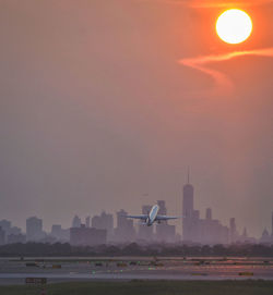 Airplane flying over buildings in city against sky during sunset
