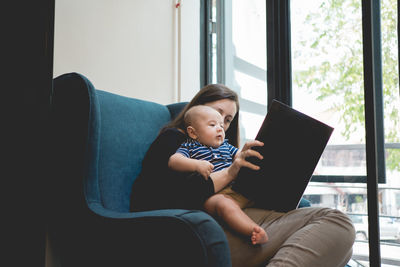 Mother with baby sitting on chair by window at home