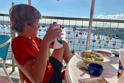 Side view of young boy having food at patio looking at a a lake 