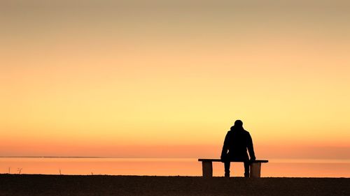 Silhouette man sitting on beach against sky during sunset