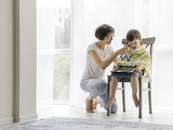 Mother cuts her son's hair by herself. little boy sits, covered with cloth, holds digital tablet.