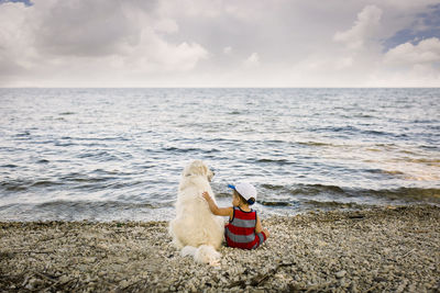 Toddler boy sitting with golden retriever dog on beach looking at lake