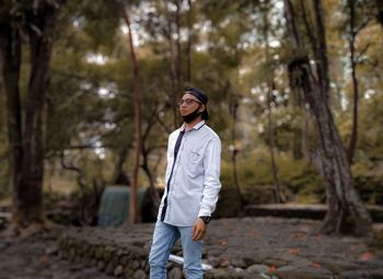 Young man looking away while standing in forest against trees