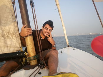 Portrait of young man gesturing peace sign while sitting in sailboat over sea