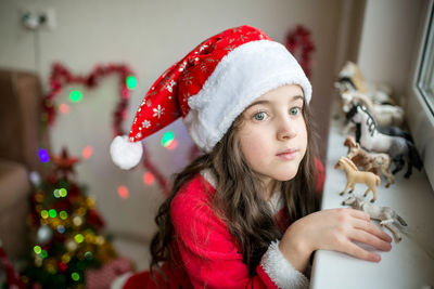 Little girl in a red dress and red christmas santa hat looking at the window