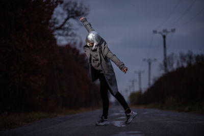 Full length of woman with arms outstretched standing on road against sky at dusk