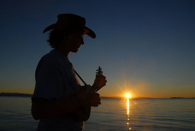 Woman playing ukulele by sea against sky during sunset