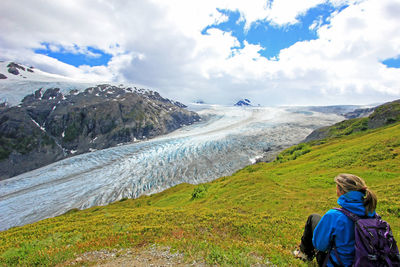 Rear view of woman sitting on grass by frozen river against sky