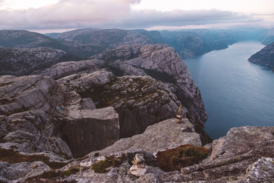 View of norwegian fjords in cloudy day, blue camping tent and unrecognizable person on red jacket standing on cliff.