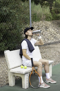 Young man sitting on a bench on a tennis court drinking water from a reusable bottle resting