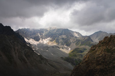 Dramatic view of forested high rocky slopes and snowy mountains in gray cloudy sky in changeable