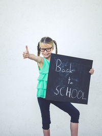 Portrait of girl gesturing thumbs up sign while holding blackboard with text against wall