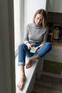 Full length of woman listening music while sitting by window