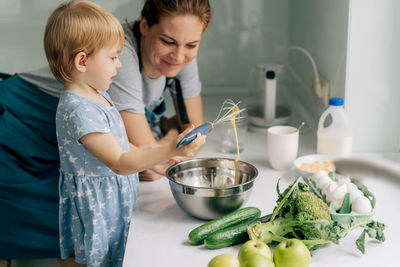 Toddler little daughter helps her mother beat eggs for an omelet.