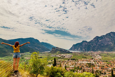 Rear view of woman with arms outstretched looking at town against cloudy sky