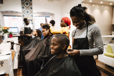 Smiling female barber making locs of male customer in salon