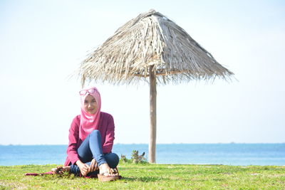 Portrait of young woman sitting on grass against thatched roof parasol at beach