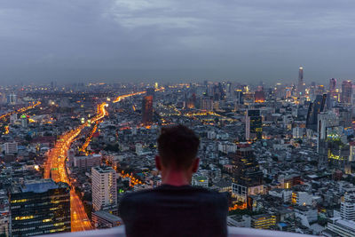 Rear view of man standing in balcony against illuminated cityscape