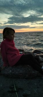 Woman sitting on beach looking at sea against sky during sunset