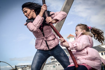 Mother pushing daughter sitting on swing in playground against sky