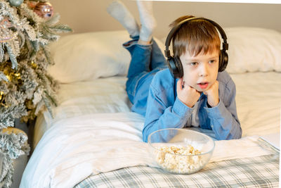 Cute boy with bluetooth headphones eating popcorn while lying in bed and watching tv