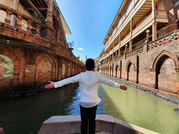 Rear view of man with arms outstretched standing on boat in canal amidst buildings