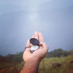 Cropped hand of man holding pocket watch against sky