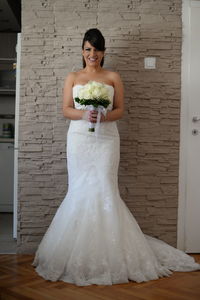 Full length of bride holding bouquet while standing against wall at home