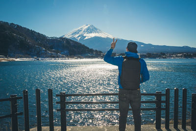 Rear view of man showing peace sign by lake against mt fuji in winter