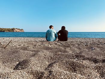 Rear view of men sitting on beach against clear sky