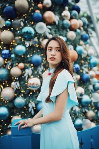 Smiling young woman looking away while standing by christmas tree outdoors