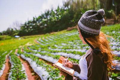 Woman wearing knit hat writing in book while standing on field