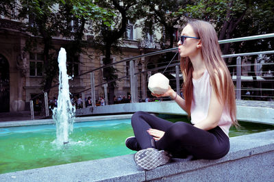 Young woman drinking coconut water while sitting by fountain