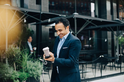 Smiling mature businessman holding disposable coffee cup with smart phone and in-ear headphones in city