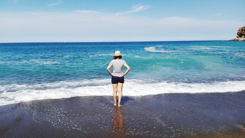 Rear view full length of woman standing with hand on hip at beach