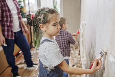 Side view of siblings working while standing against wall by father at home