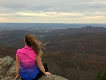 Rear view of woman looking at landscape while sitting on mountain against cloudy sky