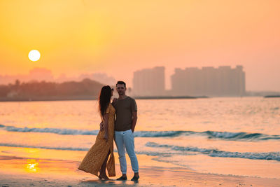 Couple standing on shore against sky during sunset
