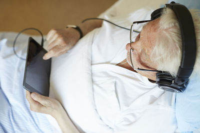 High angle view of senior man wearing headphones while touching digital tablet in hospital ward