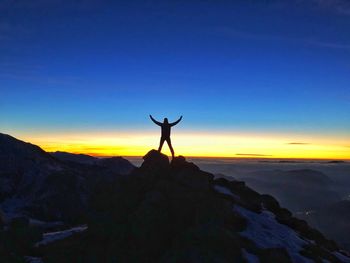 Silhouette of man standing with arms outstretched on rock against sky during sunset