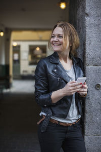 Happy woman holding mobile phone while leaning on wall outdoors