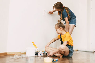 A girl with her sister in bright blue and yellow clothes helps to paint the walls in her room white.