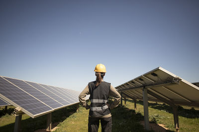 Rear view of female engineer examining solar panels while standing with arms akimbo at power station