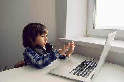 Boy child sits at a table near the window with a laptop and communicates with relatives