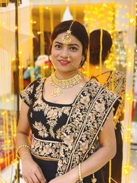 Portrait of smiling beautiful wedding guest wearing traditional clothing and jewelries against illuminated lighting equipment