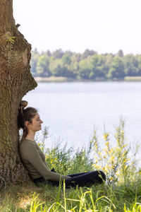 Side view of woman sitting against a tree by lake