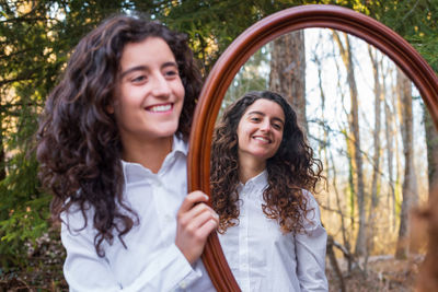 Smiling woman holding mirror with sister reflection at forest