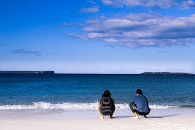 Rear view of men crouching at beach against clear blue sky with some clouds